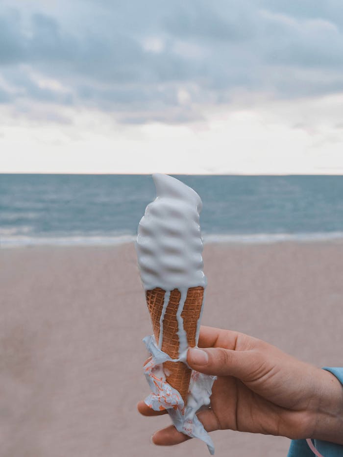 A hand holding a melting vanilla ice cream cone by the ocean under a cloudy sky.